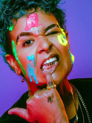 Person with neon paint on face, showing grillz on teeth, winking, and holding pinky finger to chin.