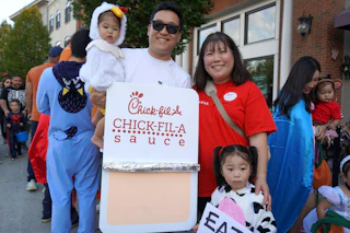 Family in costumes, including a Chick-fil-A sauce packet and a child dressed as a cow, stand outdoors at an event.