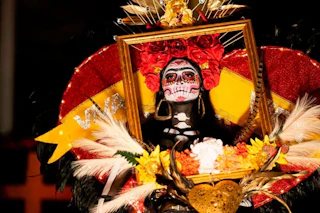 Person in Day of the Dead costume with face paint, flowers, and a golden frame around their face.