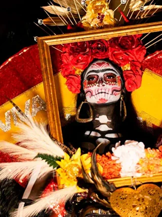 Person with Day of the Dead face paint, red flowers, and ornate costume holding frame and decorative items.