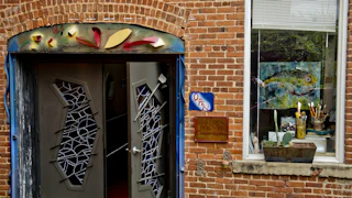 Entrance to a vibrant art studio with colorful “OPEN” sign, unique doors, and a window showcasing paintings and art supplies.