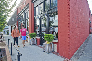 A couple walks past a red brick storefront on a sunny day in a downtown area.