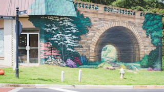 A mural depicting a stone arch bridge surrounded by lush greenery and colorful flowers on the side of a building.