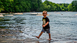 A young boy playfully stands in the Chattahoochee River water, smiling brightly under the warm sun.