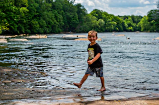 A young boy playfully stands in the Chattahoochee River water, smiling brightly under the warm sun.