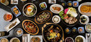 A colorful spread of Korean dishes including bibimbap, grilled meat, kimchi, and various side dishes arranged on a dark table.