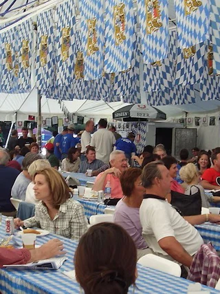 People sit at long tables in a tent decorated with blue and white flags, enjoying a festive meal together.