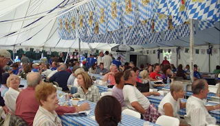 People sit at long tables in a tent decorated with blue and white flags, enjoying a festive meal together.