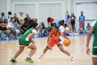 Two girls play basketball; one in orange dribbles while the other in green defends. Spectators watch.