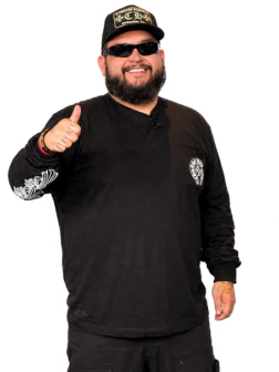 Man in black shirt, hat, and sunglasses smiling and giving a thumbs up against a white background.