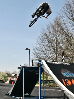 BMX rider performing an aerial trick above a ramp on a sunny day, with trees and buildings in the background.