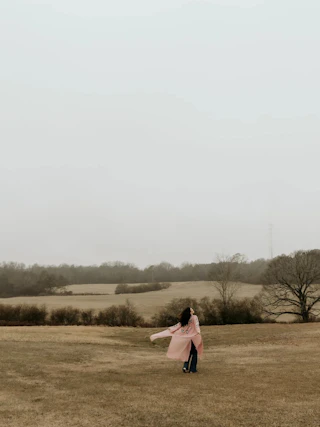 Person in a pink coat stands in an open, grassy field with trees and overcast sky in the background.