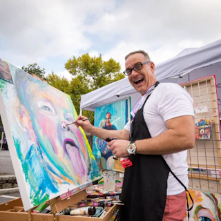 Smiling artist paints a colorful face portrait outdoors, surrounded by art supplies and a tent in the background.