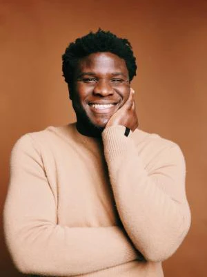 Man in a beige sweater smiling with hand on his face, standing against a brown background.