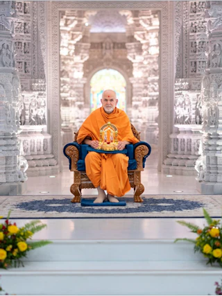 A monk in orange robes sits on a chair inside an ornate white marble temple, with flowers in the foreground.
