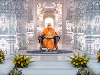 A monk in orange robes sits on a chair inside an ornate white marble temple, with flowers in the foreground.