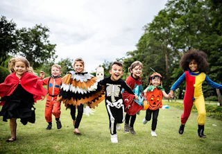 Seven children in colorful costumes run and smile outdoors on a grassy field, holding hands.