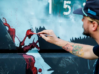 A person with a rose tattoo paints a red streak on a wall or canvas while wearing a dark cap.