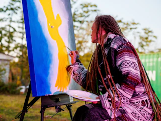 Person with long dreadlocks paints a yellow bird on a canvas outdoors, holding a plate as a palette.