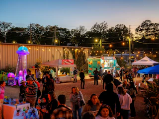 Outdoor night market with people, food trucks, tents, string lights, and a glowing jellyfish decoration.