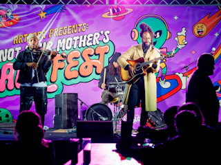 Three musicians perform on stage at Not Your Mothers Art Fest with colorful space-themed backdrop.