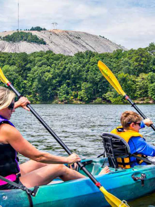Two people kayak on a lake, paddling near trees with a large rocky hill visible in the background.
