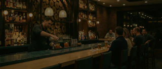 Bartender making drinks behind a bar as people sit and talk, with shelves of bottles in the background.
